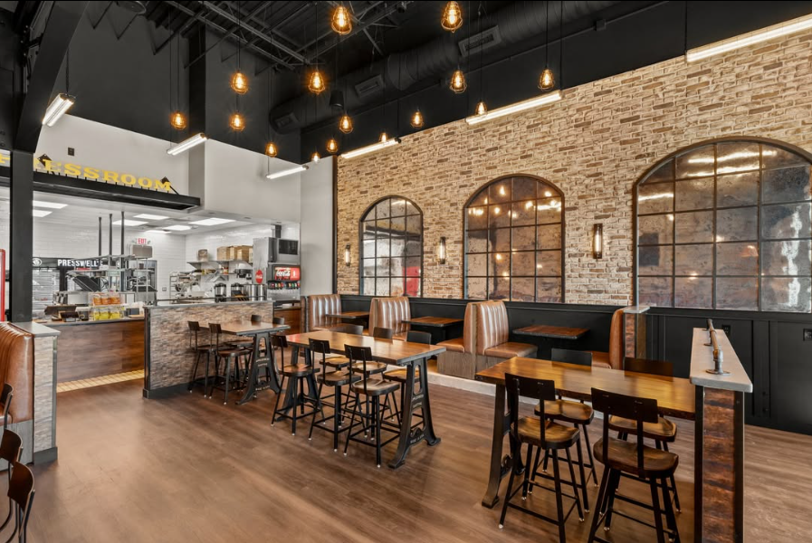 Sandwich shop dining room with wood-topped high-top tables, brown leather booths along a brick accent wall, three black arched mirrors, and the order counter on the left